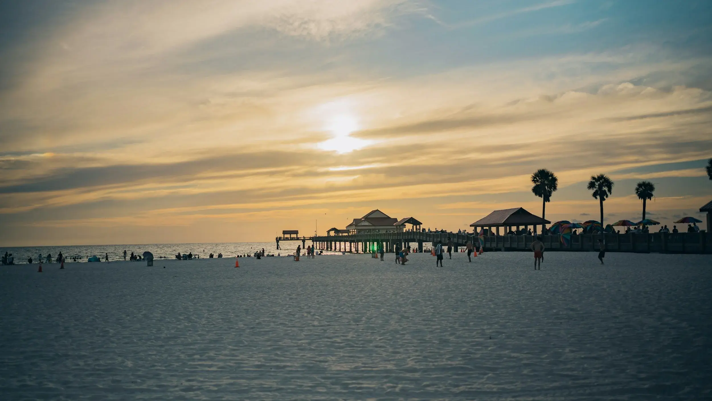 Sunset over Clearwater Beach, with a pier, palm trees, and people on the sand.